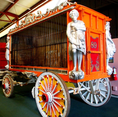 [Red and orange wagon with black bars on front and silver wooden figures at each corner. Wooden wheels are red, yellow, and white.]